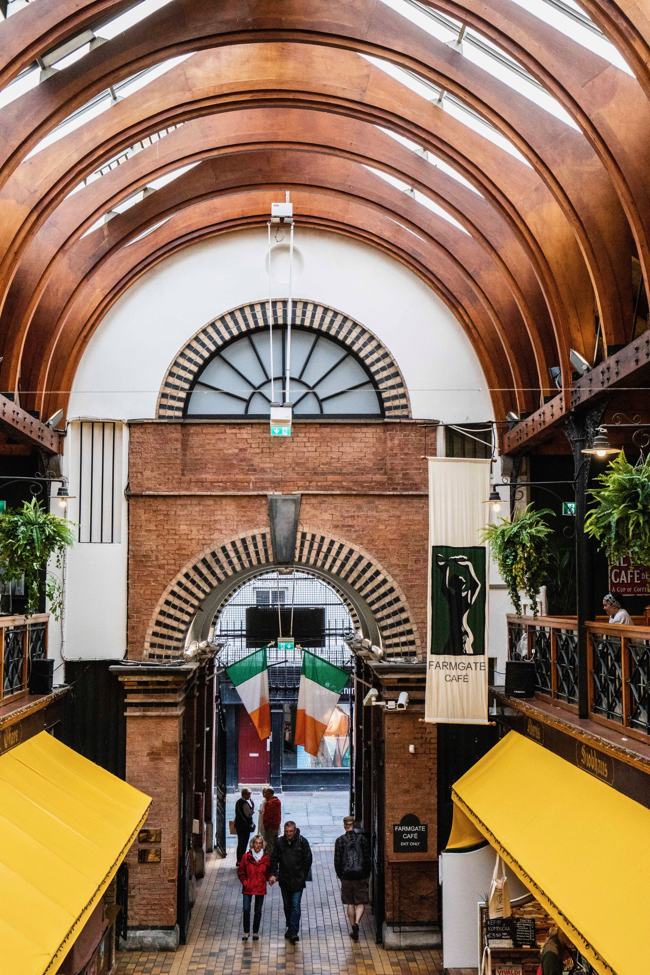 View through the arched interior of Cork&rsquo;s English Market, with the Farmgate Caf&eacute; sign, Irish flags and yellow market awnings below.